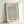 May include: A framed botanical print displaying intricate sage green and cream willow leaf patterns in an elegant repeating design. The artwork is mounted in a light wood frame with white matting, featuring "WILLOW" at the top and "WILLIAM MORRIS" at the bottom in bold black text, with "1874" in smaller print. The frame leans against a white wall, casting a soft shadow, showcasing the delicate Arts and Crafts style wallpaper pattern with flowing organic forms.