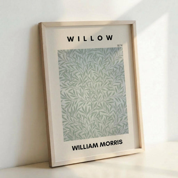 May include: A framed botanical print displaying intricate sage green and cream willow leaf patterns in an elegant repeating design. The artwork is mounted in a light wood frame with white matting, featuring "WILLOW" at the top and "WILLIAM MORRIS" at the bottom in bold black text, with "1874" in smaller print. The frame leans against a white wall, casting a soft shadow, showcasing the delicate Arts and Crafts style wallpaper pattern with flowing organic forms.