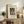 May include: Framed art print featuring a woman in historical dress holding a wicker basket, displayed on weathered wood sideboard alongside fern plant, books, ceramic vases and table lamp. The poster shows earthy tones with museum-style text layout in light wooden frame against cream matting.