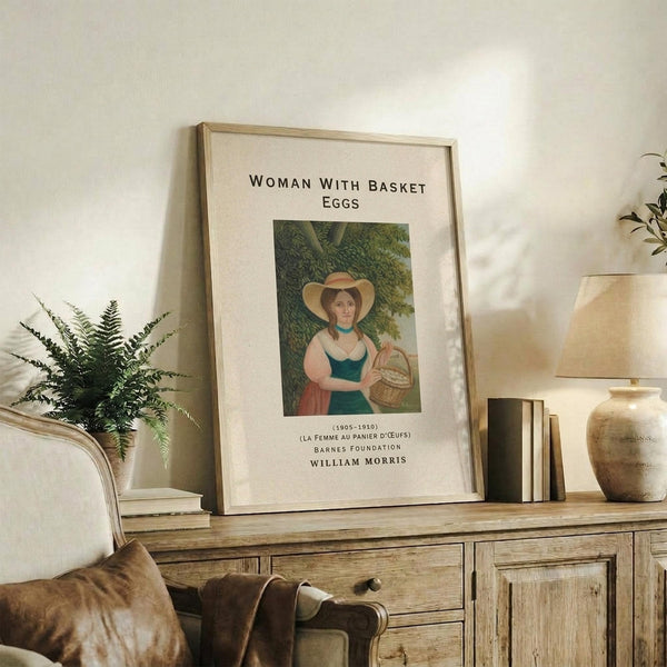May include: Framed art print featuring a woman in historical dress holding a wicker basket, displayed on weathered wood sideboard alongside fern plant, books, ceramic vases and table lamp. The poster shows earthy tones with museum-style text layout in light wooden frame against cream matting.