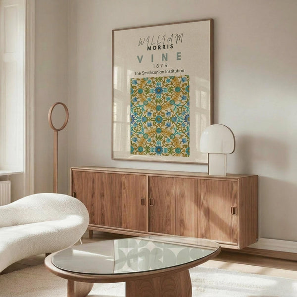 May include: William Morris botanical poster with blue and green vine pattern displayed above wooden sideboard in minimalist living room. Framed print shows intricate floral design with turquoise and olive colors. Scene includes cream upholstered chair, glass-top coffee table, and modern brass lamp on credenza.