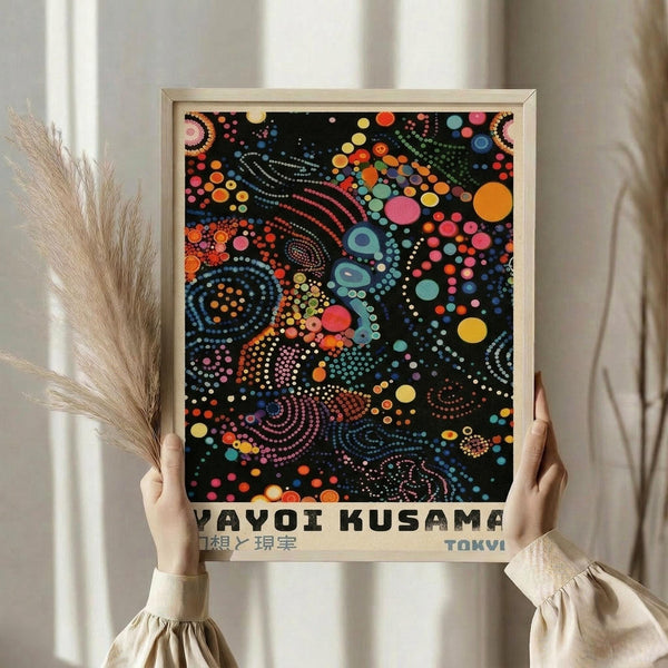 May include: Hands holding a framed art print featuring Yayoi Kusama's colorful dot painting on black background with swirling patterns in vibrant blues, oranges, pinks, and yellows. Print shows "YAYOI KUSAMA" and "TOKYO" text below artwork. Person wearing cream ruffled sleeves holds wooden-framed poster against white curtained window with dried pampas grass visible to the side.