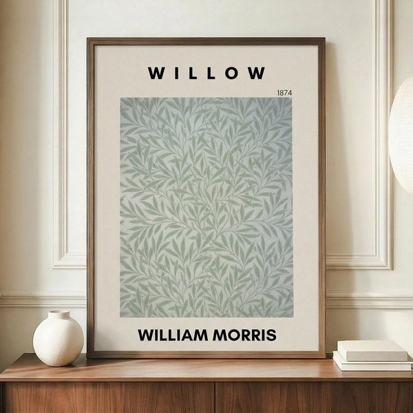May include: Framed botanical art print featuring sage green willow leaf pattern on cream background, displayed on wooden sideboard beside white ceramic vase and stacked books in elegant room with cream walls and white wainscoting