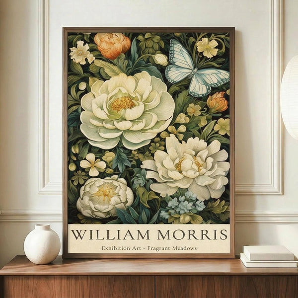 May include: Framed botanical art print displayed on wooden sideboard featuring large cream peonies, blue butterfly, and smaller flowers on dark background with William Morris exhibition text below. White ceramic vase and books sit nearby against beige paneled wall.
