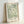 May include: Framed botanical art print with flowing green foliage pattern on cream background, displayed in natural wood frame leaning against white wall. The design features sweeping, curved leaves and organic motifs in sage green tones with "William Morris" and "Exhibition Art - Floral Reverie" text at bottom.