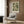 May include: Framed botanical print with flowing green foliage pattern in cream and sage colors, displayed on wood-paneled wall above beige upholstered armchair with geometric throw pillow. Modern brass floor lamp and dark side table with open book nearby. Neutral-toned contemporary living room setting with natural lighting from window.