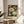 May include: Framed botanical art poster featuring white peonies, orange flowers, and blue butterfly on dark background, displayed on round wooden side table next to beige upholstered chair. Natural lighting creates soft shadows on neutral wall. Green plant leaves visible in corner.