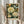 May include: Person in cream ruffled sleeves holding framed botanical art print featuring large white peonies, orange flowers, blue butterfly, and green foliage on dark background. Light wood frame, dried pampas grass visible to left, neutral curtained background with soft natural lighting.