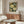May include: Framed botanical poster with white flowers, orange roses, blue butterfly on dark background displayed above wooden sideboard in modern living room with cream chair, glass coffee table, and warm natural lighting