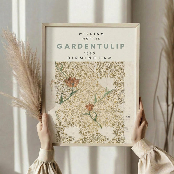 May include: Hands holding a framed botanical print featuring ornate tulip pattern in muted green and rust colors against cream background. Light wood frame, neutral interior setting with dried pampas grass and soft natural lighting through sheer curtains.