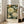 May include: Framed botanical art poster featuring cream and coral peonies, blue butterfly, and small wildflowers on dark green background, displayed in light wood frame leaning against white wall next to beige upholstered chair in bright room with large windows.