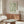 May include: Framed botanical poster with flowing green leaf pattern displayed above wooden sideboard in modern living room. Shows cream-colored chair, glass coffee table, floor lamp, and table lamp. Natural light creates soft shadows on neutral walls. Mid-century modern furniture styling with warm wood tones throughout scene.