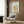 May include: Framed botanical poster with cream background featuring orange tulips and decorative golden scrollwork pattern, displayed on wood-paneled wall above beige upholstered armchair with geometric patterned pillow, brass floor lamp, and round black side table with open book in modern neutral-toned interior setting.