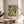 May include: Framed art print featuring flowing green and cream botanical foliage pattern in vintage tapestry style, displayed on light wood round side table next to beige upholstered chair. Soft natural lighting creates shadows on neutral wall background with plant leaves visible in foreground.