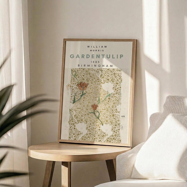 May include: Framed botanical art print displayed on round wooden side table next to beige upholstered chair. Print features green and gold floral pattern with orange tulip flowers on cream background in thin gold frame. Soft natural lighting creates shadows on white wall, with plant leaves visible in corner of neutral-toned room setting.