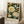 May include: Framed botanical art print leaning against white paneled wall on white fluffy rug. Features detailed illustration of cream and peach peonies, blue butterfly, and green foliage on dark background. Light wood frame, approximately 16x20 inches. White coffee cup on wooden block nearby, with beige throw blanket visible at right edge.