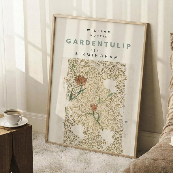 May include: Framed vintage botanical print leaning against white wall next to textured beige throw blanket. The poster features ornate green paisley pattern with red tulip flowers on cream background, displaying "WILLIAM MORRIS GARDENTULIP 1885 BIRMINGHAM" text at top. Small wooden side table with white coffee cup visible on left. Natural lighting creates soft shadows on the light-colored floor and wall.