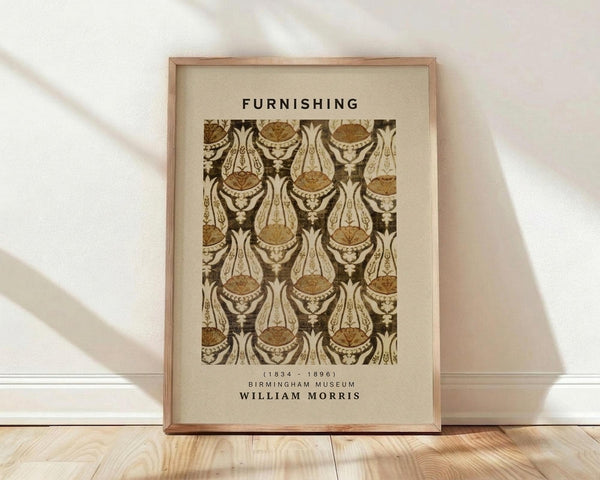 May include: Framed vintage William Morris textile pattern poster in warm brown and cream tones, featuring ornate paisley and floral motifs in repeating design. Print shows museum attribution text and artist dates. Natural wood frame leans against white wall on light wooden floor with soft natural lighting creating diagonal shadows.