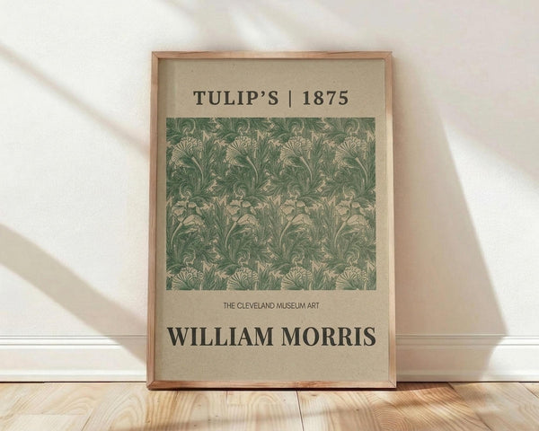 May include: Framed poster print with green and cream botanical tulip pattern from 1875, displayed in light wood frame against white wall on wooden floor. Vintage William Morris floral design with intricate leaves and blooms in sage green tones on cream background. Museum-style art print with text header and attribution.