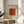 May include: Framed art print with cream background featuring rust orange geometric pattern of repeating circular motifs arranged in grid. Text reads "William Morris Smithsonian Institution 1870" and "Famous Artwork Pattern" in black lettering. Displayed on round wooden side table next to beige upholstered chair with plant leaves visible, soft natural lighting creating shadows on white wall.