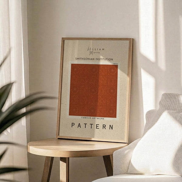 May include: Framed art print with cream background featuring rust orange geometric pattern of repeating circular motifs arranged in grid. Text reads "William Morris Smithsonian Institution 1870" and "Famous Artwork Pattern" in black lettering. Displayed on round wooden side table next to beige upholstered chair with plant leaves visible, soft natural lighting creating shadows on white wall.