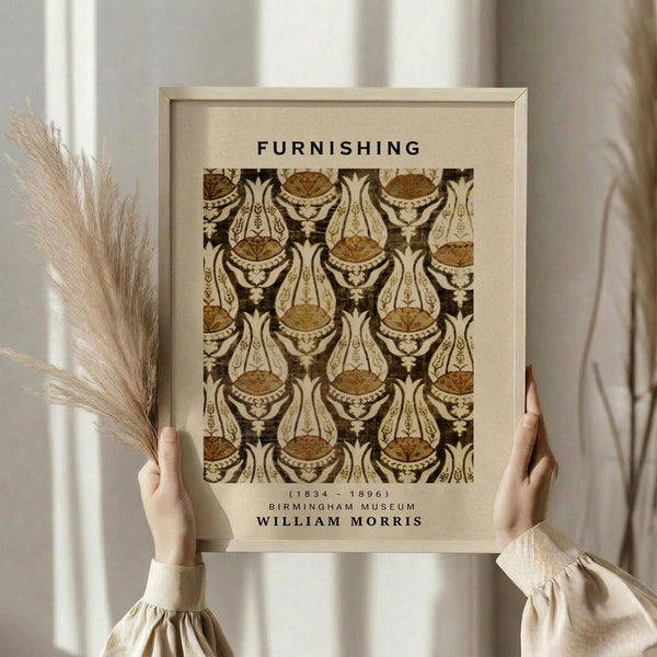 May include: Hands holding a framed vintage botanical pattern print in warm brown and cream tones, featuring ornate floral motifs in an Art Nouveau style. The wooden frame contains text reading "FURNISHING" at top and "BIRMINGHAM MUSEUM WILLIAM MORRIS" at bottom. Person wears a beige blouse and holds dried pampas grass, standing against soft natural lighting near white curtains.