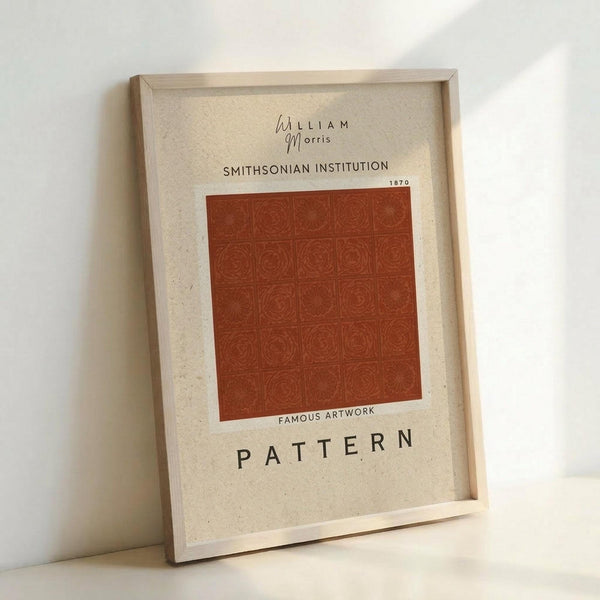 May include: Framed poster print leaning against white wall on light surface. Light wooden frame contains cream-colored paper with red-orange geometric pattern featuring repeated circular motifs in grid layout. Black text reads "William Morris" at top, "Smithsonian Institution 1870" below, "Famous Artwork" in middle, and "Pattern" at bottom. Soft natural lighting creates gentle shadows on minimalist background.