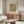 May include: Framed William Morris textile pattern poster with ornate paisley design in brown and cream tones hanging above a mid-century modern walnut credenza. White sculptural chair and glass-top coffee table visible in minimalist living room with cream walls and natural lighting.