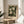 May include: Framed botanical art print displayed on a round wooden side table next to a beige upholstered chair. The artwork features white and cream flowers with green foliage on a dark background, housed in a thin gold frame with cream matting. Soft natural light creates shadows across the neutral-toned room setting with a plant visible in the corner.