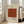 May include: Framed vintage-style poster with beige background featuring William Morris and Smithsonian Institution text above a rust-colored square filled with repeating circular botanical pattern. Large "PATTERN" text below. Light wood frame leans against white wall next to beige upholstered chair in bright living room with large windows.