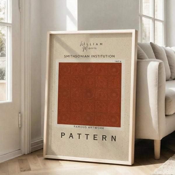 May include: Framed vintage-style poster with beige background featuring William Morris and Smithsonian Institution text above a rust-colored square filled with repeating circular botanical pattern. Large "PATTERN" text below. Light wood frame leans against white wall next to beige upholstered chair in bright living room with large windows.
