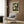 May include: Framed botanical art print featuring cream-colored flowers and dark green foliage against black background, displayed on wood-paneled wall above beige upholstered chair with geometric patterned pillow. Modern brass floor lamp and dark round side table with open book complete the neutral-toned interior setting with natural lighting from window.
