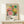 May include: Colorful vintage exhibition poster featuring vibrant polka dot patterns and radiating floral designs in pink, yellow, green and orange, displayed in natural wood frame on light wooden console table. Small wrapped gift with twine and greenery sits nearby. Poster shows museum exhibition details and Japanese characters against cream background.