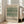May include: Framed botanical poster with vintage green tulip pattern design leaning against white wall near beige upholstered chair. Light wooden frame contains ornate floral wallpaper-style artwork in sage green and cream tones. Bright room setting with large windows, hardwood floors, and natural sunlight creating shadows on white walls.