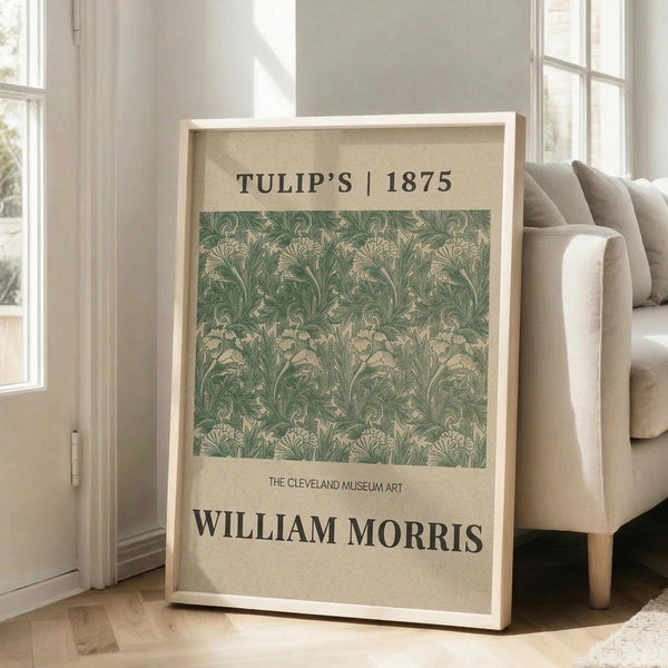 May include: Framed botanical poster with vintage green tulip pattern design leaning against white wall near beige upholstered chair. Light wooden frame contains ornate floral wallpaper-style artwork in sage green and cream tones. Bright room setting with large windows, hardwood floors, and natural sunlight creating shadows on white walls.