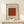 May include: Framed art print with William Morris and Smithsonian Institution text above a rust-colored geometric pattern featuring repeated circular motifs arranged in a grid. The beige background poster reads "Famous Artwork Pattern 1870" below the design. Light wood frame sits on pale wooden surface next to small wrapped gift with evergreen sprig against cream wall.