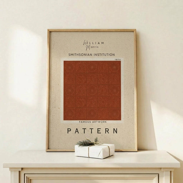 May include: Framed art print with William Morris and Smithsonian Institution text above a rust-colored geometric pattern featuring repeated circular motifs arranged in a grid. The beige background poster reads "Famous Artwork Pattern 1870" below the design. Light wood frame sits on pale wooden surface next to small wrapped gift with evergreen sprig against cream wall.