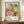 May include: Framed exhibition poster featuring vibrant circular floral patterns in pink, yellow, green, and orange with polka dot backgrounds, displayed on wooden sideboard next to white ceramic vase and stacked books against cream wainscoted wall. Poster shows colorful abstract botanical design with radiating petals and geometric shapes in Kusama's signature dotted style.