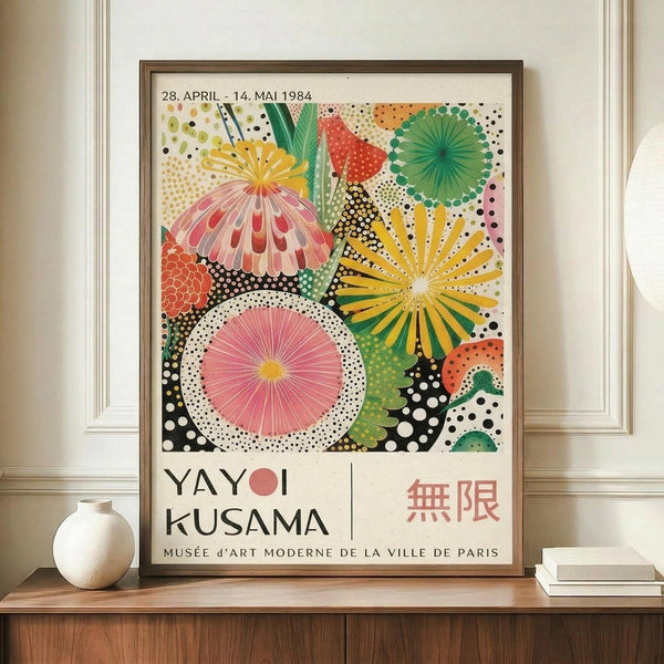 May include: Framed exhibition poster featuring vibrant circular floral patterns in pink, yellow, green, and orange with polka dot backgrounds, displayed on wooden sideboard next to white ceramic vase and stacked books against cream wainscoted wall. Poster shows colorful abstract botanical design with radiating petals and geometric shapes in Kusama's signature dotted style.