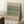 May include: Framed poster with green botanical tulip pattern on cream background, leaning against white wall corner next to coffee cup on wooden block and beige blanket on white textured rug