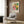 May include: Colorful Yayoi Kusama exhibition poster featuring vibrant geometric patterns, polka dots, and radial designs in pink, green, yellow, and orange, displayed in light wooden frame on warm wood paneled wall above beige upholstered armchair with geometric throw pillow, brass floor lamp, and dark round side table with open book nearby.
