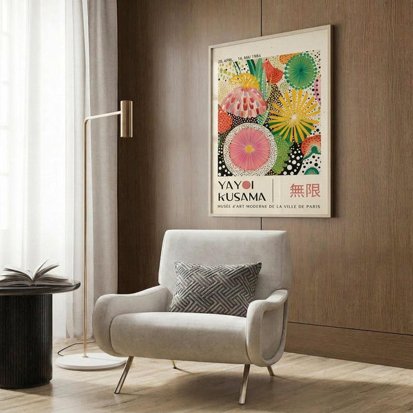 May include: Colorful Yayoi Kusama exhibition poster featuring vibrant geometric patterns, polka dots, and radial designs in pink, green, yellow, and orange, displayed in light wooden frame on warm wood paneled wall above beige upholstered armchair with geometric throw pillow, brass floor lamp, and dark round side table with open book nearby.