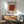 May include: Framed poster with William Morris pattern design in rust orange tones displayed above wooden sideboard. Mid-century modern living room setting with sage green armchair, white table lamp, and round coffee table. Natural sunlight creates geometric shadows on white wall.