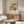 May include: Colorful Yayoi Kusama exhibition poster with vibrant circular patterns in pink, yellow, green, and orange displayed above a mid-century modern wooden credenza. The artwork features radiating dots and geometric designs in a contemporary living room with white curved chair, glass coffee table, and minimalist decor.