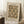May include: Framed vintage-style poster with "FURNISHING" text and ornate brown and cream decorative pattern featuring stylized vessels and flourishes, attributed to William Morris (1834-1896) Birmingham Museum. Light wood frame leaning against white paneled wall beside wooden side table with white coffee cup, cream textured rug, and soft beige throw blanket visible.