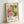 Colorful exhibition poster featuring vibrant circular and radiating patterns in pink, yellow, green, and orange with polka dot backgrounds. The artwork displays geometric flowers and sunburst designs against black dotted areas. Text shows dates "28. APRIL - 14. MAI 1984" at top, "YAYOI KUSAMA" in large letters, Japanese characters, and "MUSÉE d'ART MODERNE DE LA VILLE DE PARIS" at bottom. The poster is displayed in a light wooden frame leaning against a white wall.