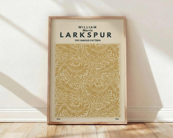May include: Framed poster print featuring William Morris Larkspur pattern in golden yellow on cream background, displayed against white wall on light wooden floor. The intricate botanical design shows flowing leaves and floral motifs in Art Nouveau style. Natural wood frame complements the warm tones. Soft natural lighting creates gentle shadows on the wall and floor.