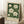 May include: Framed botanical art print leaning against white wall corner, featuring white magnolia flowers with green leaves on dark emerald background, cream matting and light wood frame, positioned next to coffee cup on wooden block and beige knit throw on white textured rug