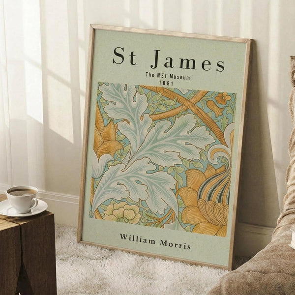 May include: Framed poster with sage green background featuring ornate botanical pattern in golden yellow and pale green. Shows decorative leaves, flowers and curved Art Nouveau-style motifs. Text reads "St James The MET Museum 1881" and "William Morris" in black serif font. Natural wood frame leans against white wall corner next to white coffee cup on wooden block and cream textured throw blanket on white fuzzy rug.