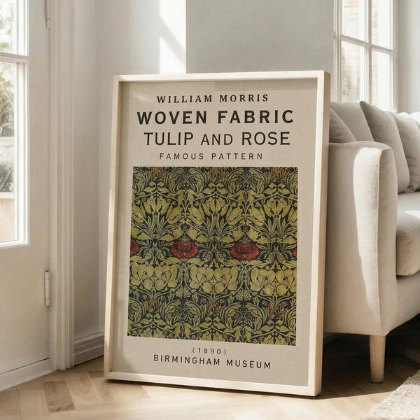 May include: Framed poster displaying William Morris woven fabric pattern with tulips and roses in olive green, burgundy red, and golden yellow tones against cream background. Light wood frame leans against white wall next to beige upholstered sofa in bright, modern living room with large windows and natural lighting.